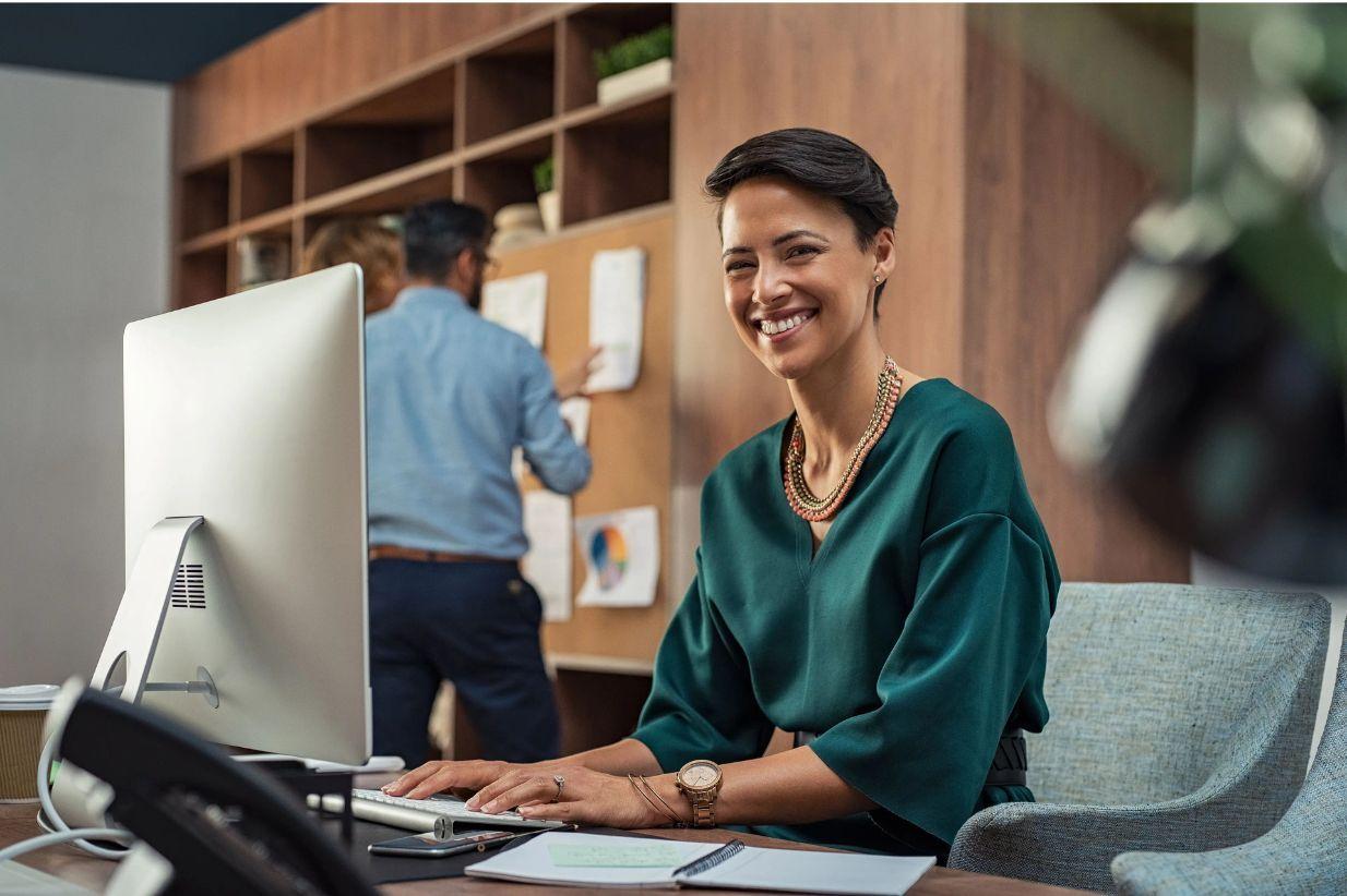 Smiling woman working on computer in a modern office, a colleague in the background.