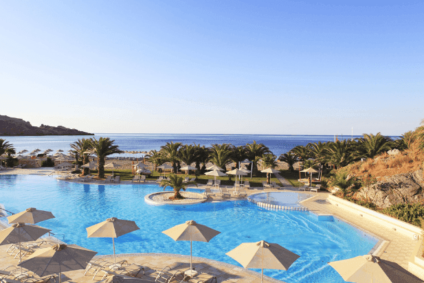 Large blue swimming pool with umbrellas overlooking a sandy beach and the clear sea.